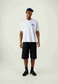 Balta kokvilnas T-kreklu ar grafisku dizainu uz krūtīm, kombinēta ar tumšiem džinsa šortiem. Aksesuāri ietver zilu cepuri un saulesbrilles.