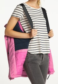 Osoba w szarych dżinsach i białej koszuli w paski, trzymająca ręką paski od czarnego plecaka, z różową kurtką zarzuconą na ramionach.