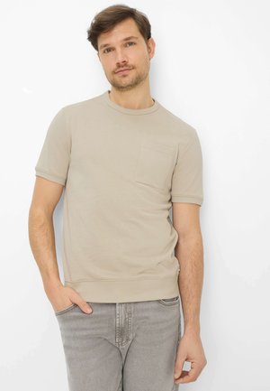 Uomo con capelli castani corti che indossa una camicia beige a maniche corte con tasca, in piedi con una mano nella tasca dei jeans grigi contro sfondo bianco.