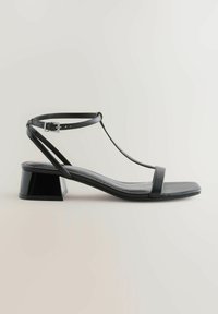 Sandalia de charol negro con un diseño de tira en T, punta cuadrada y un tacón bajo. Cuenta con una correa en el tobillo ajustable para un ajuste seguro.