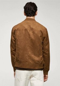 Veste bomber en daim marron avec une finition texturée, des poignets et un ourlet côtelés, et un dos épuré. Associée à un pantalon de couleur claire.