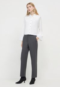 Blouse blanche courte avec fermeture à boutons, associée à un pantalon gris ajusté et des bottines noires à talons pointus. Tissu doux, coupe structurée.