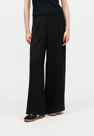 Άτομο που φοράει φαρδιά μαύρη παντελόνα, μαύρο τοπ και μαύρα αθλητικά παπούτσια με καφέ σόλες, στέκεται μπροστά σε ένα απλό ανοιχτό φόντο.