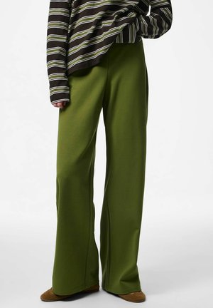 Personne portant un pantalon ample vert olive, des chaussures marron, et un haut à manches longues rayé avec des rayures horizontales vertes, blanches et noires.