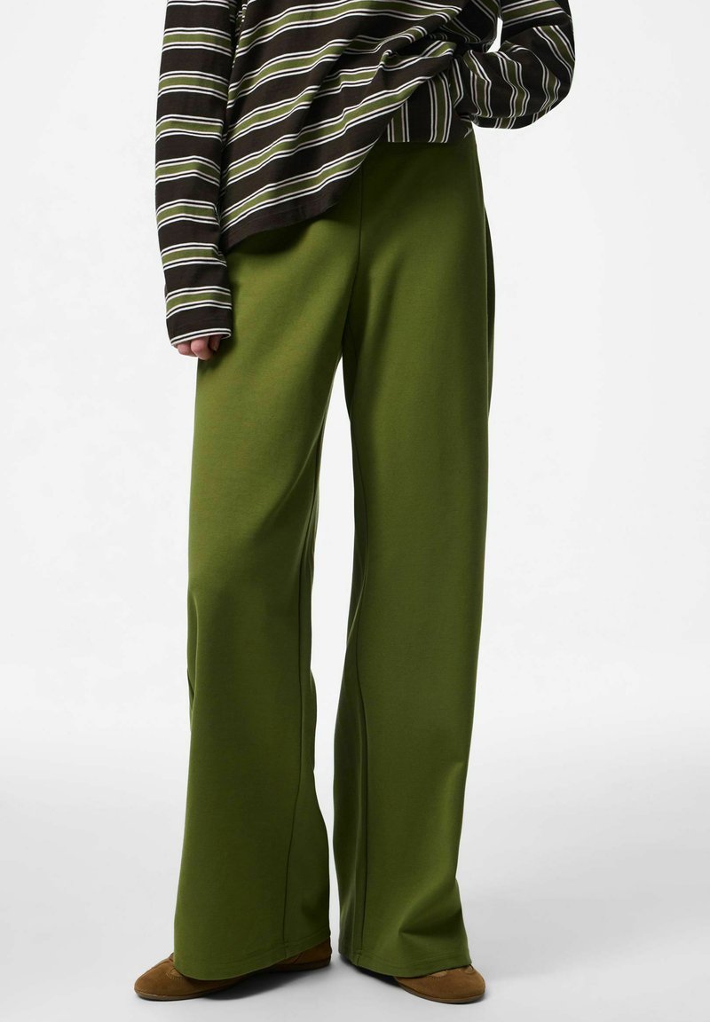 Personne portant un pantalon ample vert olive, des chaussures marron, et un haut à manches longues rayé avec des rayures horizontales vertes, blanches et noires.