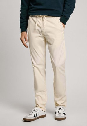 Mann trägt beige Kordhose, dunkelgrünes Sweatshirt und weiße Sneaker mit schwarzen Akzenten, steht mit einer Hand in der Tasche.