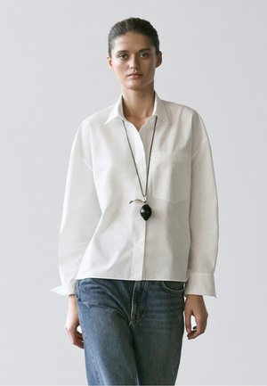 Jeune femme portant une chemise blanche à col, un jean bleu et un collier pendentif noir sur un fond clair uni.