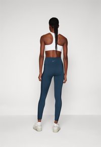 Magas derekú, sötétkék leggings sima textúrával, hátul logóval, fehér sportmelltartóval és világos lábbelivel párosítva.