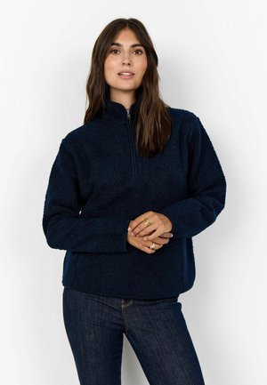 Navy fleece pullover met een half-zip kraag, gestructureerd oppervlak en lange mouwen. Draagt in combinatie met donkere jeans, wat een ontspannen pasvorm laat zien.