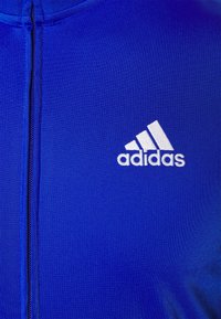 Vêtement bleu à zip avec logo Adidas blanc sur la poitrine gauche, comportant trois bandes diagonales au-dessus du nom de la marque.