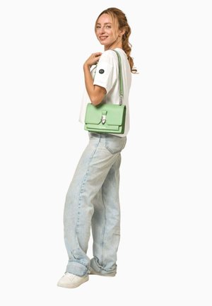 Jeune femme portant un t-shirt blanc, un jean bleu clair et des baskets blanches, tenant un sac à bandoulière vert clair avec une lanière chaîne argentée.
