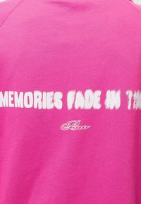 Sudadera rosa con la frase "LOS RECUERDOS SE DESVANECEN CON EL TIEMPO" en blanco, con una textura suave y un pequeño logo cerca del dobladillo.