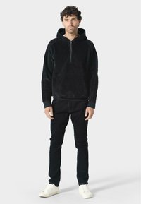 Pullover nero con cappuccio realizzato in tessuto texturizzato, con zip frontale a metà e tasca frontale. Abbinato a jeans neri e sneakers bianche.