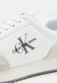Zapatilla blanca con una parte superior sintética lisa, acentos de ante y un logo gris que presenta "Calvin Klein Jeans". Cordonera plana y suela de goma.
