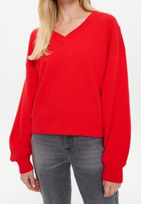 Vrouw in een rode V-hals lange mouwtrui met oversized mouwen en grijs gewassen jeans tegen een effen achtergrond.