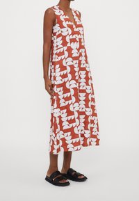 Röd ärmlös klänning med ett geometriskt mönster av vita blad, med en v-ringning och når till mellankalven, tillsammans med svarta sandaler.