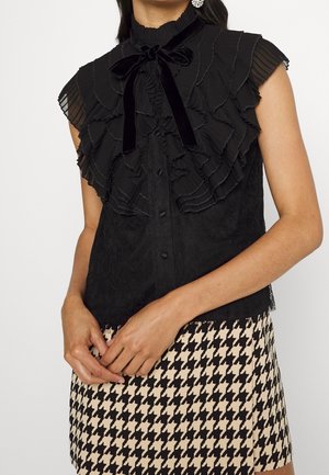 Kvinde iført en sort flæset ærmeløs bluse med en fløjs sløjfe og et beige-og-sort hundetandsmønstret skørt, hænderne foldet foran.