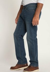 Jeans in denim blu scuro con taglio dritto, dotati di patta con cerniera, cinque tasche e un leggero sbiadimento. Indossati con scarpe di pelle marroni.