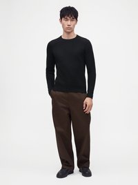 Homme debout portant un pull noir à manches longues, un pantalon marron à jambes larges et des chaussures noires sur un fond blanc uni.