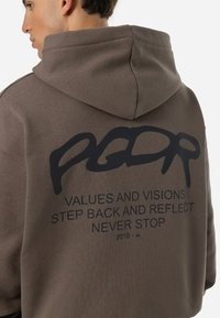 Sudadera marrón con un acabado texturizado, que presenta un gran gráfico negro en la parte trasera con texto. Incluye capucha con cordón y un ajuste relajado.