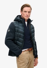 Giacca da uomo in materiale doppio con parte superiore in piumino trapuntato blu scuro e corpo in lana blu navy testurizzato. Include cappuccio e dettaglio del logo.