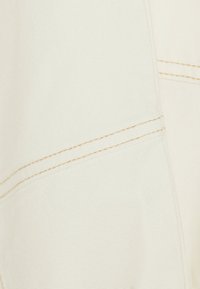 White fabric with close-up view of beige double stitching detail forming diagonal and vertical lines.