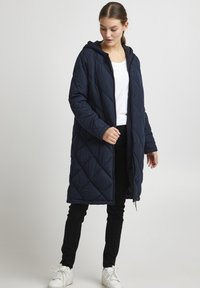Long navy quilted puffer coat with a hood, featuring a front zipper, diamond pattern, and worn over a white top and black pants. White sneakers.