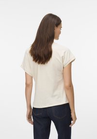 Camiseta de manga corta color crema con puños enrollados, textura suave y corte relajado, usada con jeans azul oscuro, vista desde atrás.