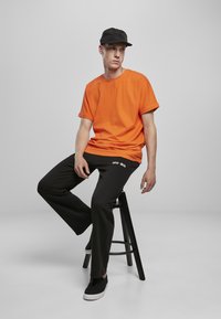 Tricou portocalie cu mânecă scurtă, pantaloni negri casual, șapcă neagră și adidași negri cu talpă albă, așezat pe un scaun negru, pe un fundal gri.
