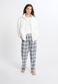 Weiße Kunstfell-Jacke mit Reißverschluss und langen Ärmeln, kombiniert mit schwarz-weißen karierten Schlafanzughosen mit elastischem Bund.