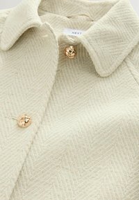 Crème herringbone textuurjas met gouden gebraid knopen en een puntige kraag, merklabel "NEXT" zichtbaar aan de binnenkant van de hals.