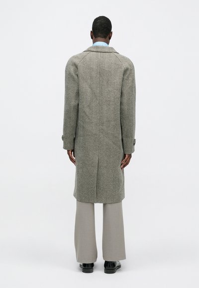 Sivi kaput u riblji kostim s definiranim oblikom, s oštrim ovratnikom i dvostrukim kopčanjem sprijeda, uparen s sivim hlačama.