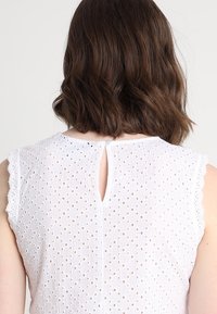 Bílý bezrukávový top s klíčovou dírou na zádech, jemnou krajkovou vpředu na ramenech a texturovaným vzorem s otvory po celé ploše.