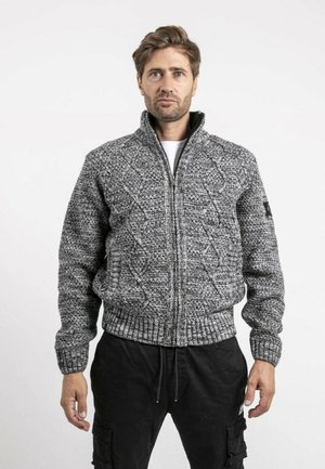 Homme portant un pull zippé gris à torsades et un pantalon cargo noir, debout devant un fond blanc uni.