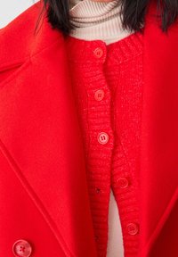 Manteau en mélange de laine rouge avec des revers crantés, associé à un cardigan en rib rouge avec des boutons assortis. Les deux vêtements présentent une finition texturée.