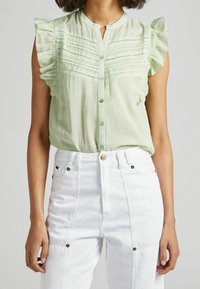 Jasnozielona, bezrękawnikowa bluzka z falbaniastymi ramionami, z zapięciem na guziki i poziomymi teksturowanymi paskami. Zestawiona z białymi spodniami o wysokim stanie.