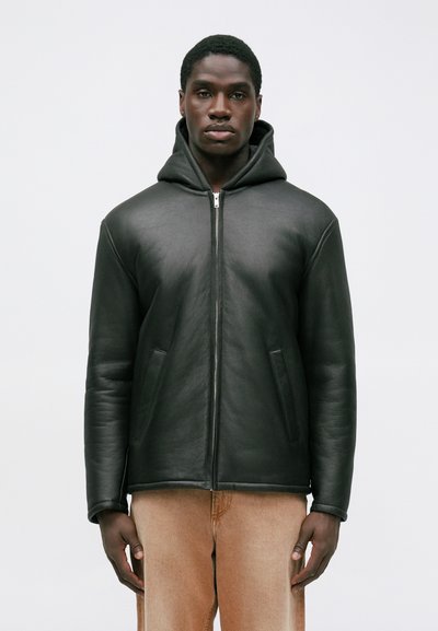 Black leather zip-up jacket with a hood, featuring side pockets and a smooth texture. Worn over light beige trousers.