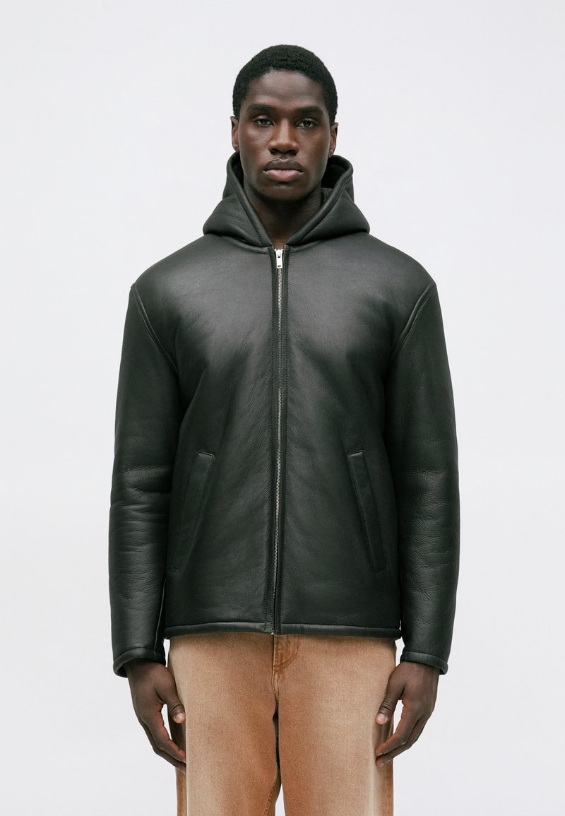 Schwarze Lederjacke mit Reißverschluss und Kapuze, mit seitlichen Taschen und glatter Textur. Über hellbeigen Hosen getragen.