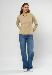 Vrouw in een beige poloshirt met lange mouwen, blauwe jeans en witte sneakers, staand met de handen achter de rug tegen een effen achtergrond.