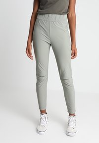 Světle šedé sportovní kalhoty s elastickým páskem, zužujícími se nohavicemi a detaily na kolenou, v kombinaci s bílými teniskami. Hladká, elastická látka.