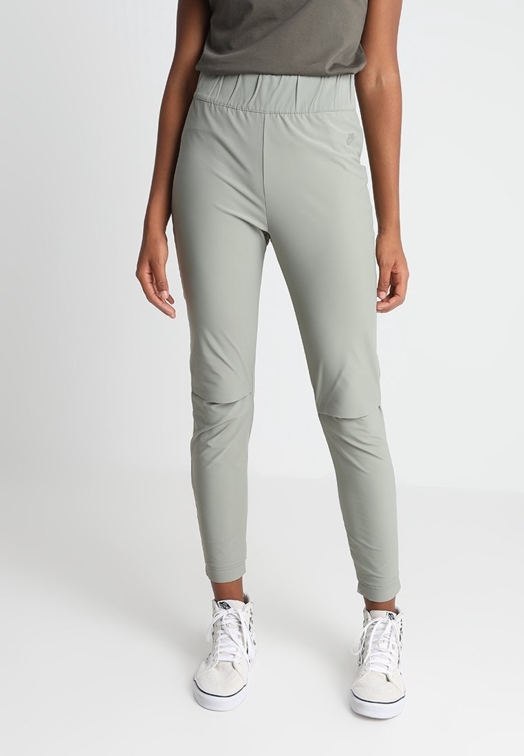 Světle šedé sportovní kalhoty s elastickým páskem, zužujícími se nohavicemi a detaily na kolenou, v kombinaci s bílými teniskami. Hladká, elastická látka.