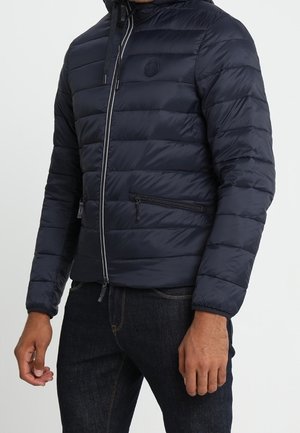 Mężczyzna ubrany w granatową pikowaną kurtkę z zamkiem błyskawicznym z przodu i zamykanymi kieszeniami, w połączeniu z ciemnymi jeansami.