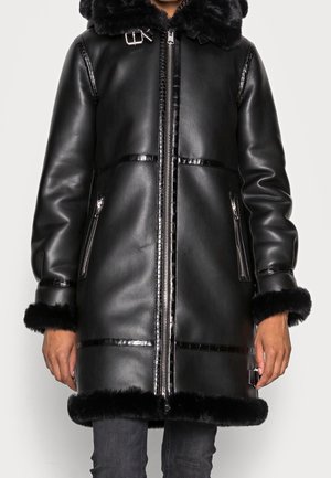 Manteau en cuir synthétique noir avec une capuche noire en fourrure. Présente des accents brillants, des zips sur les côtés et des bordures en fourrure sur les manches et l'ourlet.