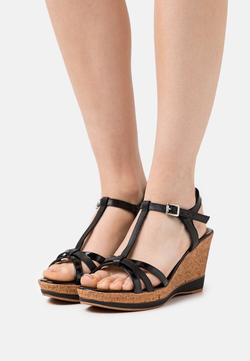 Sandalias de cuña de charol negro con suelas de corcho. Cuenta con una correa en el tobillo ajustable, diseño de punta abierta y tiras cruzadas en la parte frontal.