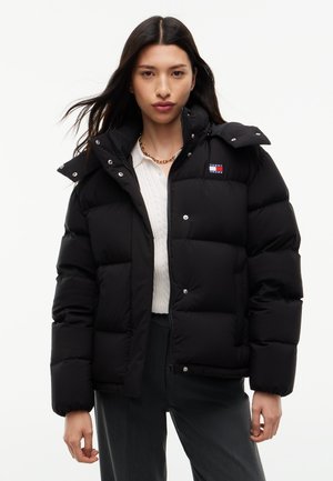 Kvinde iført en sort dunjakke med høj krave, hvid sweater, mørke bukser og en guldkædehalskæde mod en ensfarvet baggrund.