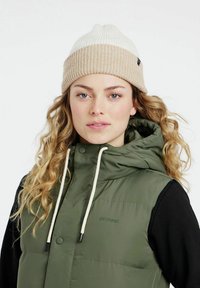 Žalias pūkinis liemenė su kapišonu, baltas ir šviesiai rudas megztas beanie. Paprastas dizainas, kontrastingos tekstūros ir ryškūs raišteliai ant liemenės.