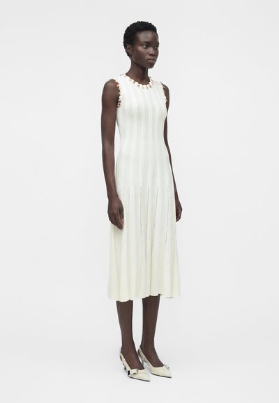 Υψηλή γυναίκα με κοντά μπούκλια φοράει αμάνικο λευκό midi φόρεμα με υφή ρίγες και μπεζ χαμηλοτάκουνα παπούτσια, στέκεται πάνω σε λευκό φόντο.