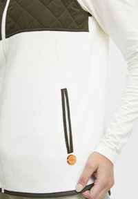Vit fleecejacka med en kviltad olivgrön övre del, med svarta detaljer och en trälogoknapp vid fållen.