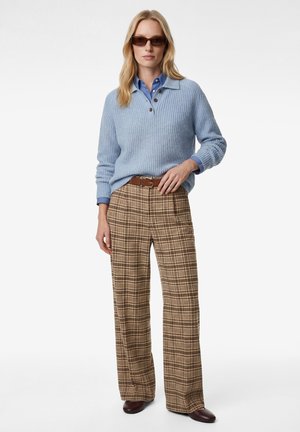 Femme portant un pantalon large à carreaux marron, une chemise bleue à col sous un pull tricoté bleu clair, une ceinture marron, des lunettes de soleil et des chaussures marron.