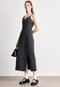 Vestido midi de denim preto com decote em V, alças finas, racha frontal e tachas decorativas ao longo do corpete. Modelo segura uma pequena bolsa preta.